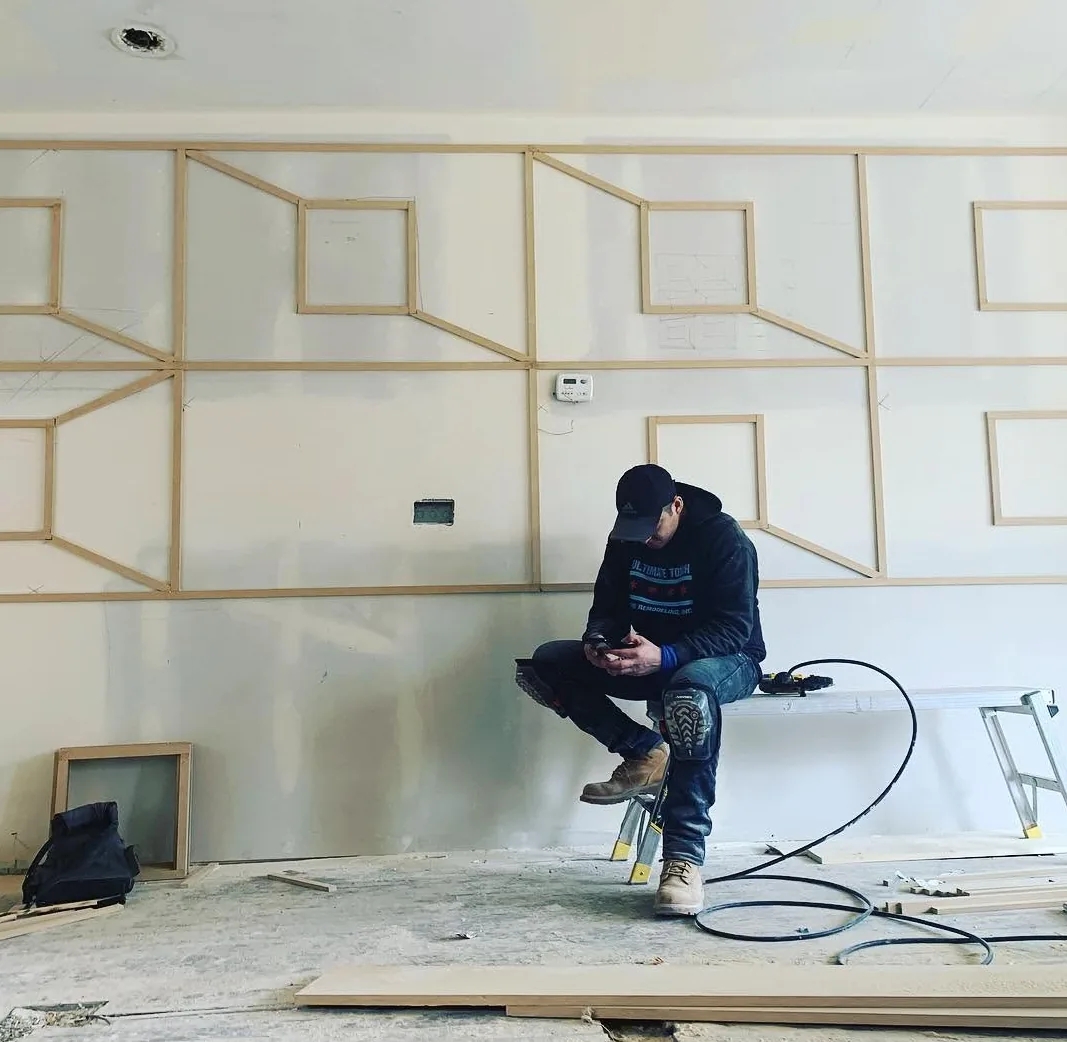 An Ultimate Touch carpenter fitting geometric picture-frame trim to a drywalled accent wall, with milled molding stacked on the floor