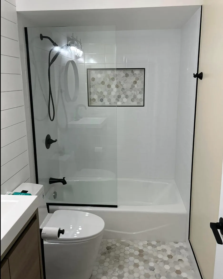 Finished bathroom remodel with a white-tiled tub and shower, matte black fixtures, a hexagon mosaic niche, matching hex-tile floor and a frameless glass panel