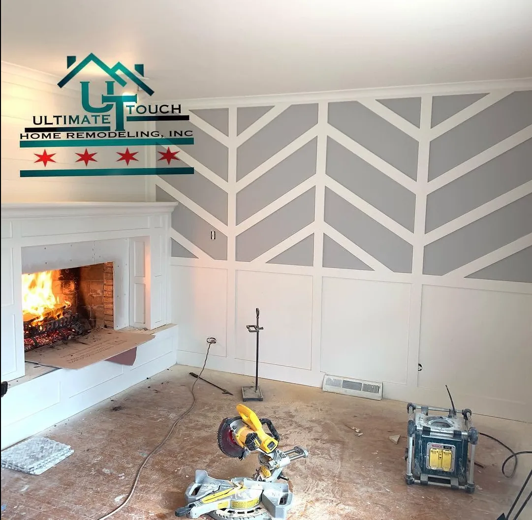 A chevron-patterned trim feature wall and white board-and-batten fireplace surround under construction, with a lit firebox and a miter saw on the subfloor